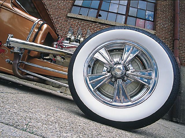 Chrome wide-whitewall wheel on a bronze hot rod, brick building background
